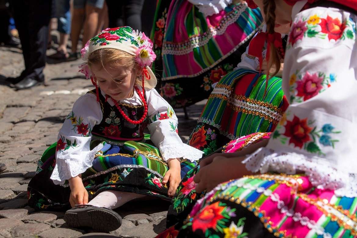 Een jong meisje in kleurrijke traditionele folkloristische kleding zit op kasseien en past haar schoen. Andere mensen in vergelijkbare geborduurde outfits zijn in de buurt en het zonlicht accentueert hun levendige patronen.