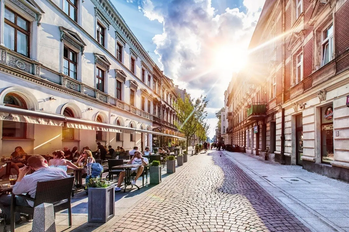 Mensen zitten aan de buitentafels van een café aan een geplaveide straat met historische gebouwen, terwijl het zonlicht door een gedeeltelijk bewolkte hemel stroomt en een warme en levendige sfeer creëert.