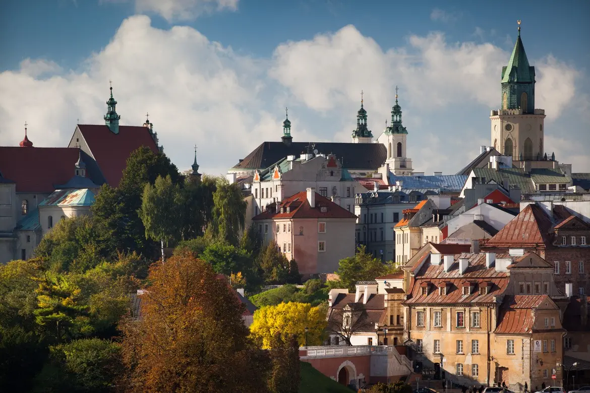 Een schilderachtig uitzicht op een historische Europese stad met kleurrijke gebouwen, kerktorens en groene bomen onder een gedeeltelijk bewolkte hemel, die een herfstachtige omgeving suggereert.