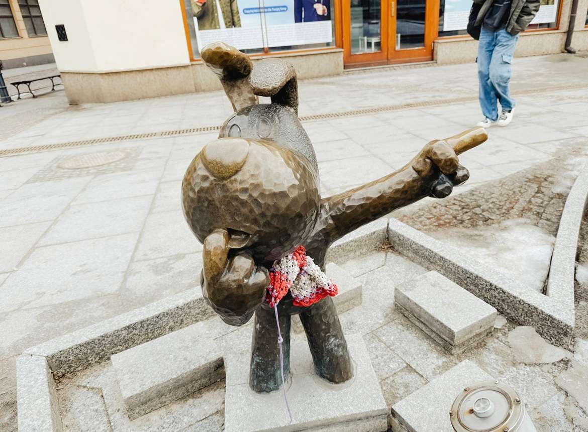 Een bronzen beeld van een cartooneske hond met een opgeheven en wijzende arm, die een roodwitte sjaal draagt, staat op een stenen platform op een trottoir in de stad. Op de achtergrond is een persoon te zien.