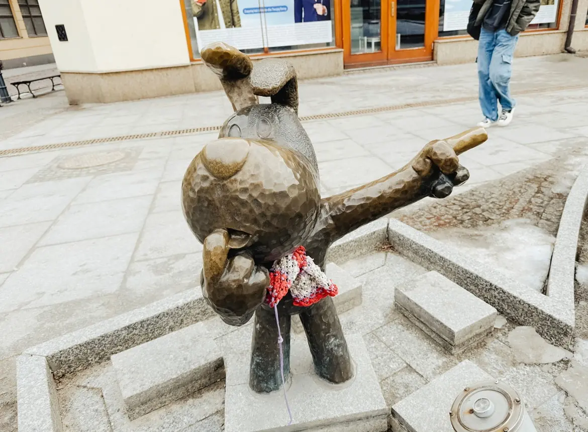 Een bronzen beeld van een cartooneske hond met een opgeheven en wijzende arm, die een roodwitte sjaal draagt, staat op een stenen platform op een trottoir in de stad. Op de achtergrond is een persoon te zien.